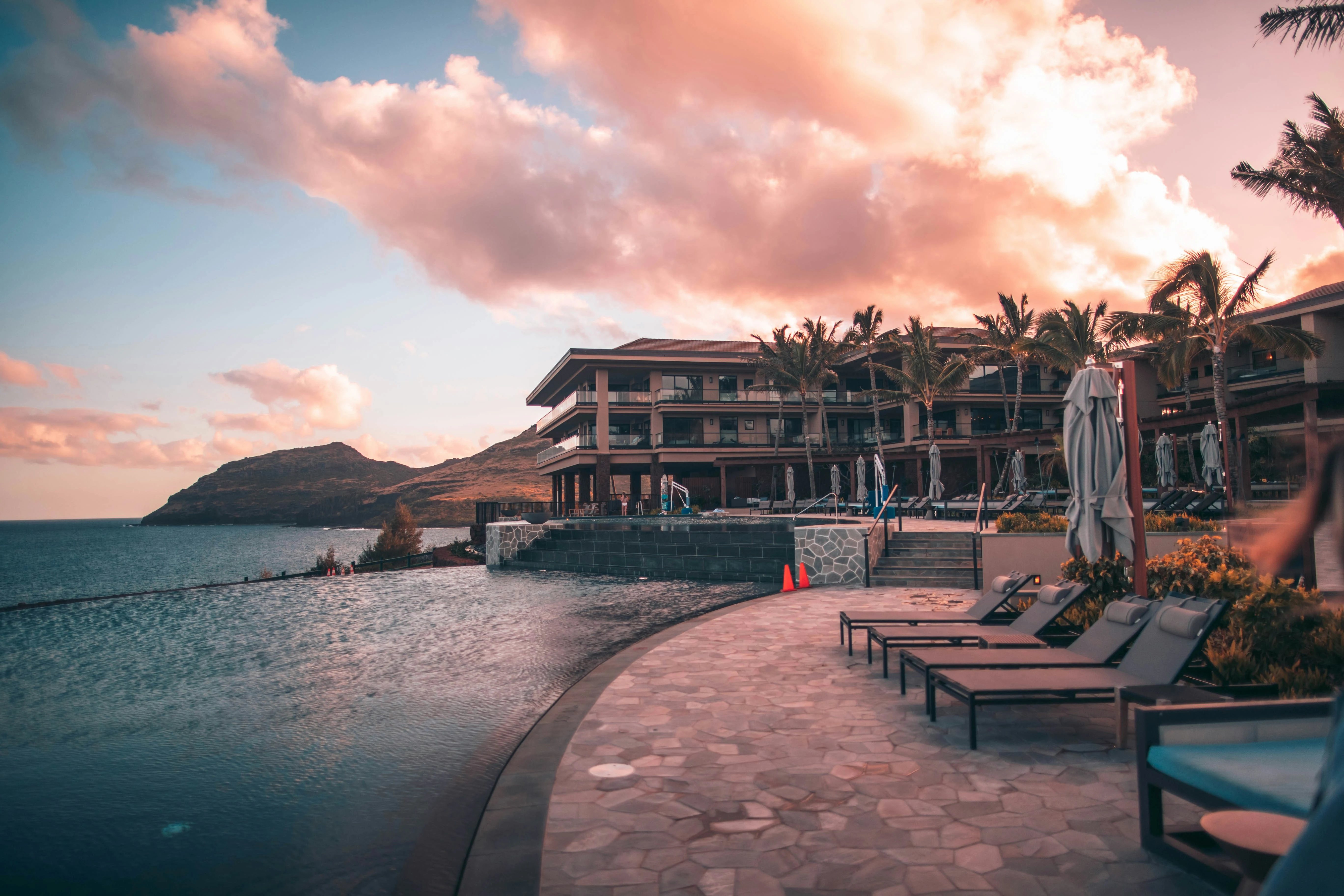 Coastal luxury resort infinity pool and sun loungers at sunset against dramatic coastal hills